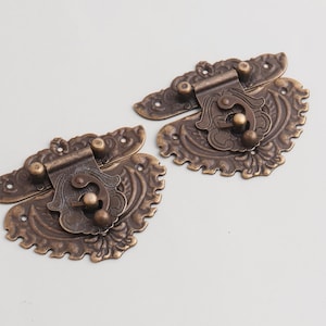 以下が含まれることがあります： 2つのアンティークブロンズ色のラッチで、華やかなフローラルデザインが施されています。各ラッチには装飾的な留め金と、取り付け用の複数のネジ穴があります。金属はヴィンテージで、古びた外観をしています。