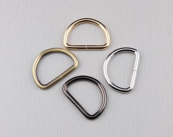 May include: Four metal D-rings in gold, antique brass, silver, and black. The D-rings are arranged in a circular pattern, each with a visible seam. The background is a light gray, textured surface.