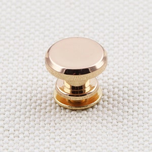 May include: A close-up of a gold-colored metal button. The button has a rounded top and a cylindrical base with a smaller, circular bottom. The button is highly reflective, with a polished finish.