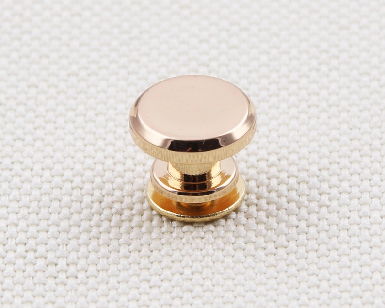 May include: A close-up of a gold-colored metal button. The button has a rounded top and a cylindrical base with a smaller, circular bottom. The button is highly reflective, with a polished finish.