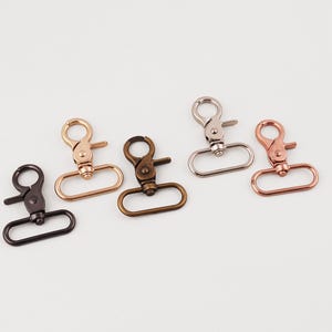May include: An assortment of metal swivel clasps in various finishes, including black, gold, bronze, silver, and rose gold. Each clasp features a spring-loaded gate and an oval-shaped ring for attaching to straps or other hardware. Ideal for crafting and DIY projects.