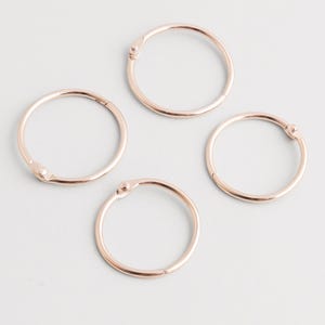 Puede incluir: Cuatro anillas de encuadernación de oro rosa dispuestas sobre una superficie blanca. Cada anilla es circular con una abertura articulada. Las anillas son de un material metálico brillante, ideales para organizar documentos.