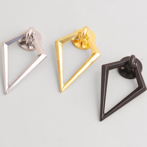 Op de afbeelding: Drie geometrische ladegrepen in zilver, goud en donker brons. Elke greep heeft een ruitvormige handgreep die aan een cirkelvormige basis is bevestigd. Deze grepen zijn ontworpen voor meubels en kasten.