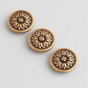 May include: Three antique brass-colored cabinet knobs. Each round knob features a detailed floral design in the center, surrounded by a circular border. The knobs are arranged diagonally on a light gray background.