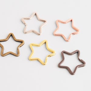 May include: Five star-shaped metal clasps in various metallic finishes: bronze, rose gold, gold, silver, and dark bronze. The clasps are open and ready to be attached to a keychain or other craft project.
