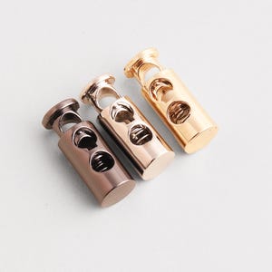 Puede incluir: Tres cierres de cordón metálicos en bronce, oro rosa y oro. Cada cierre de cordón tiene un cuerpo cilíndrico con dos agujeros y un mecanismo de resorte en el interior. La parte superior presenta un lazo para sujetar un cordón.