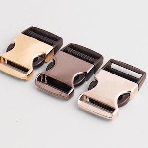 Puede incluir: Tres hebillas metálicas de liberación lateral en colores oro, bronce y plata. Cada hebilla tiene un componente de plástico negro y un diseño curvo. Las hebillas están dispuestas sobre un fondo blanco.