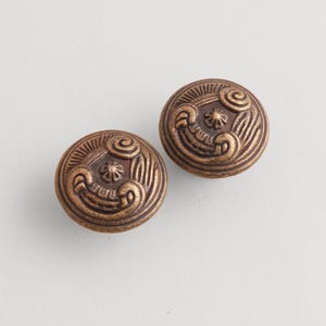 May include: Two antique bronze-colored cabinet knobs. Each round knob features a detailed, swirling design with a central star-like element. The knobs have a vintage aesthetic, suitable for furniture restoration or decorative projects.