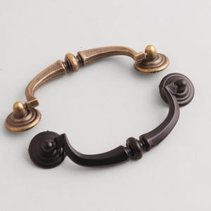 May include: Two metal drawer pulls with a curved, swinging handle design. One pull features a weathered, antique brass finish with visible highlights, while the other pull has a solid, matte black finish. Both include circular mounting plates.