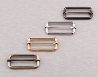 May include: Four metal rectangle sliders in different finishes: dark bronze, silver, gold, and antique brass. Each slider has a rectangular frame with a central bar. These hardware components are used in various crafting and sewing projects.