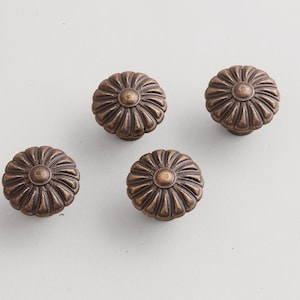 May include: Four bronze-colored cabinet knobs with a flower-like design. Each knob has a central circular element surrounded by petal-shaped ridges. The knobs are arranged on a light gray surface.