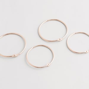May include: Four rose gold binder rings are arranged on a white surface. Each ring is circular with a small, rounded clasp. The rings are made of a shiny, metallic material and are evenly spaced apart.
