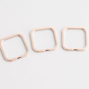 Peut inclure: Trois porte-clés carrés en or rose sont disposés en diagonale sur une surface blanche. Chaque anneau a des coins arrondis et une petite ouverture. Les porte-clés sont en métal et ont une finition brillante.