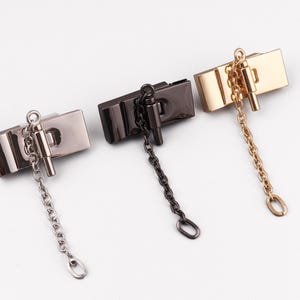 Op de afbeelding: Drie metalen portemonneesluitingen in zilver, zwart en goud. Elke sluiting is rechthoekig met een ketting en een kleine, cilindrische sluiting. De sluitingen zijn gerangschikt op een wit oppervlak.