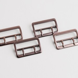 Può includere: Quattro fibbie metalliche color bronzo con un telaio rettangolare e tre barre interne. Le fibbie sono disposte su una superficie bianca, mostrando il loro design e colore.