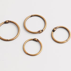 Op de afbeelding: Vier antiek messingkleurige bindringen van verschillende formaten zijn gerangschikt op een wit oppervlak. Elke ring heeft een scharnierende opening met een kleine, ronde klinknagel. De ringen zijn cirkelvormig en hebben een licht versleten uiterlijk.