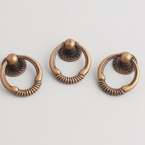 Op de afbeelding: Drie antiek bronzen kastknoppen met een ringontwerp. Elke knop heeft een cirkelvormige ring met een getextureerde grip en een decoratieve basis. De hardware is gemaakt van metaal en heeft een vintage esthetiek.
