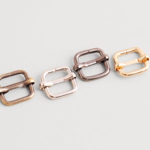Puede incluir: Cuatro hebillas correderas metálicas en acabados latón antiguo, plata, metal de cañón y oro. Cada hebilla tiene un marco cuadrado con una barra central para el ajuste de la correa. Ideal para la fabricación de bolsos y manualidades.