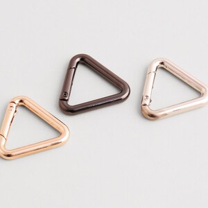 Pode incluir: Três mosquetões triangulares de metal nas cores ouro rosa, bronze e ouro. Os clipes estão dispostos sobre uma superfície cinza claro. Estes achados de hardware são usados para vários projetos de artesanato e acessórios.