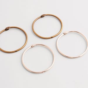 Puede incluir: Cuatro anillas metálicas para encuadernación en varios colores: bronce, dorado y oro rosa. Los anillos circulares tienen una abertura con bisagra para sujetar páginas. El fondo es gris claro.