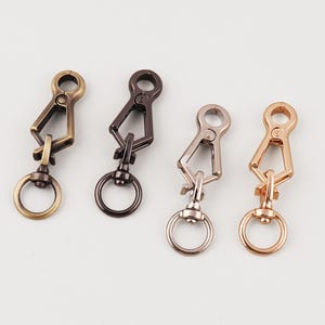 May include: Four metal swivel clasps in antique brass, dark bronze, silver, and rose gold. Each clasp has a ring at the bottom and a swivel hook at the top, suitable for attaching to keychains or bags. The clasps are arranged on a white surface.