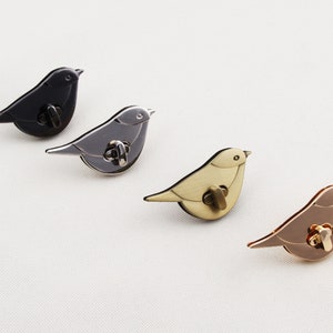 May include: Four metal bird-shaped clasps in black, grey, gold, and rose gold. Each clasp has a circular locking mechanism. The birds are arranged diagonally on a white surface, showcasing their different finishes and designs.