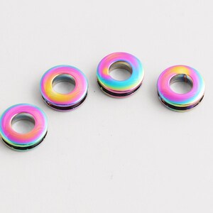 Könnte beinhalten: Vier irisierende Metallperlen in Donutform. Die Perlen haben einen Regenbogeneffekt mit Farbtönen von Rosa, Gelb, Grün und Blau. Die Perlen sind auf einer weißen Oberfläche angeordnet.