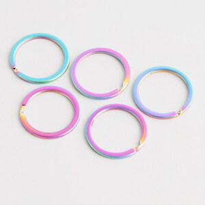 May include: Five iridescent key rings in shades of blue, pink, and yellow lie on a white surface. The rings are circular with a hinged opening. These colourful rings are suitable for keychains or craft projects.