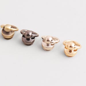 May include: Four metal bag hardware pieces in various finishes: antique brass, gunmetal, rose gold, and gold. Each piece features a circular ring attached to a rounded base, suitable for bag closures or decorative accents.