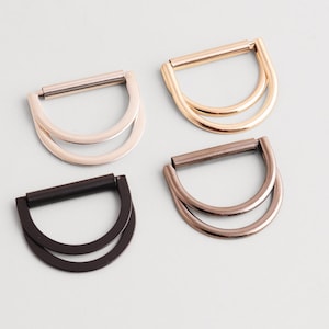 May include: Four metal D-ring buckles in various finishes: silver, gold, bronze, and black. Each buckle has a D-shaped design with a double-loop structure, suitable for bag making or other craft projects.