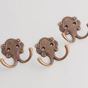 May include: Three antique bronze-colored metal hooks. Each hook has a decorative, swirling design on the backplate and a curved hook. The hooks are approximately 3 inches wide and 2 inches tall.