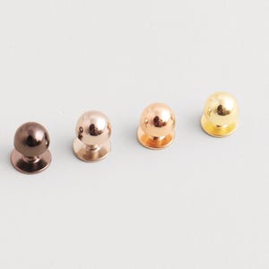 Puede incluir: Cuatro gemelos metálicos en fila, cada uno con una parte superior redondeada y una base plana. Los colores son bronce oscuro, oro rosa, cobre y oro. Los gemelos están sobre una superficie gris claro.