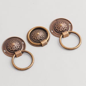 May include: Three antique bronze cabinet pulls with a floral design. Each pull features a circular backplate with a detailed flower and a ring handle. The pulls are made of metal and have a vintage aesthetic.