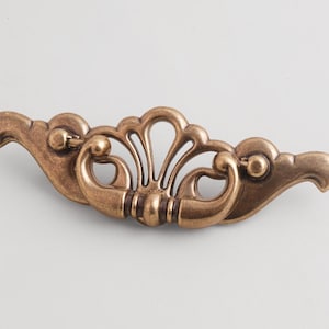 May include: A horizontal drawer pull handle made of antique brass-toned metal. The hardware features a central fan-shaped motif with symmetrical curved arms extending outward, ending in rounded, bird-like head shapes on both sides.