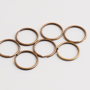 Peut inclure: Sept porte-clés de couleur bronze sont disposés sur une surface blanche. Chaque anneau est circulaire avec une petite ouverture. Les anneaux ont une finition métallique et sont espacés uniformément.