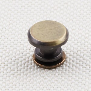 May include: A small, antique-style cabinet knob with a round, brass-colored top and a dark, bronze-colored base. The knob is designed for easy gripping and is likely used for drawers or cabinet doors. The hardware is set against a neutral background.