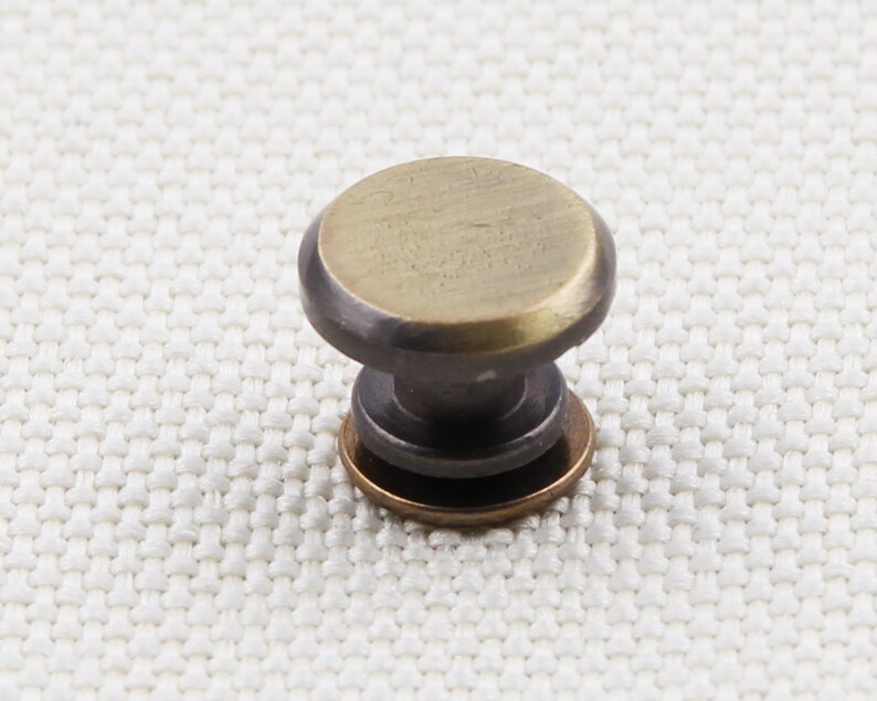 May include: A small, antique-style cabinet knob with a round, brass-colored top and a dark, bronze-colored base. The knob is designed for easy gripping and is likely used for drawers or cabinet doors. The hardware is set against a neutral background.