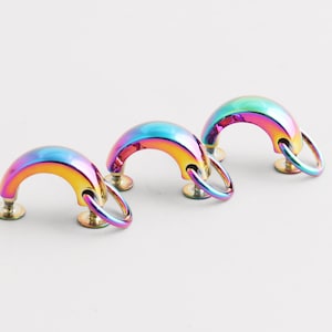 Könnte beinhalten: Drei schillernde, regenbogenfarbene Schrankgriffe. Jeder Griff hat ein gebogenes, halbkreisförmiges Design mit einem angebrachten Ring. Die Griffe sind auf kleinen, runden, goldfarbenen Sockeln montiert. Die Griffe sind wahrscheinlich für Möbel.