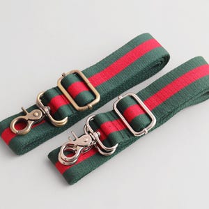 May include: Two green and red striped bag straps with metal hardware. One strap has gold-toned clasps and a rectangular buckle, while the other has silver-toned clasps and a rectangular buckle. The straps are likely for a purse or handbag.