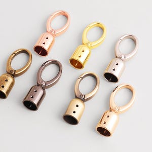 May include: An assortment of metal swivel clasps in various finishes, including gold, bronze, rose gold, and gunmetal. Each clasp features a bell-shaped base with two small holes and an oval-shaped ring for attachment. The clasps are arranged on a light gray surface.