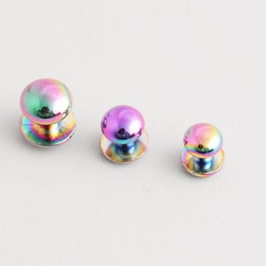 May include: Three iridescent metal studs with a round, domed top and a flat, circular base. The studs reflect a rainbow of colours, including pink, green, and gold. The studs are of varying sizes.