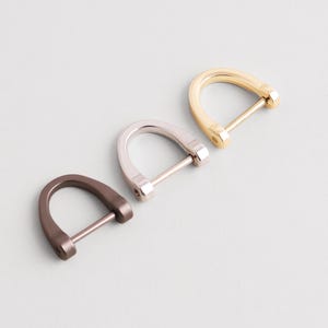 Op de afbeelding: Drie metalen D-ringgespen in verschillende afwerkingen: brons, zilver en goud. De D-ringen zijn gebogen met een rechte staaf aan de onderkant. Deze fournituren worden gebruikt bij het maken van tassen en leerbewerking.
