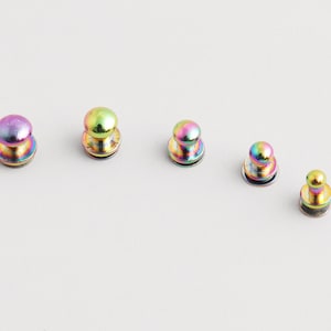 May include: Five iridescent metal button studs are displayed. Each stud has a rounded top and a flat base. The colours shift from purple to green and gold. The studs are arranged on a white surface.