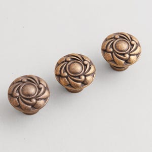 Puede incluir: Tres tiradores de armario de bronce antiguo con diseño floral. Los tiradores son redondos con un patrón en espiral en relieve alrededor de un círculo central. El acabado tiene un aspecto cálido y envejecido, lo que sugiere un estilo vintage o clásico.