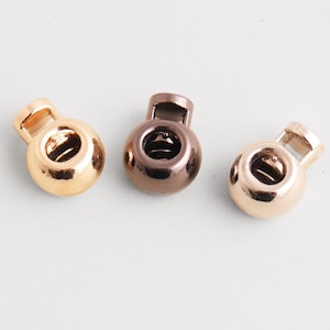 Puede incluir: Tres cierres de cordón metálicos en tonos dorados y bronce se disponen sobre una superficie blanca. Cada cierre de cordón tiene un cuerpo redondeado con un agujero y una abertura rectangular en la parte superior. Se utilizan para proyectos de costura y manualidades.