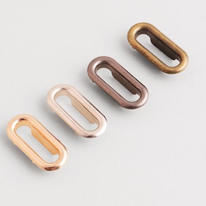 Könnte beinhalten: Vier ovale Metallösen in verschiedenen Metallic-Ausführungen: Gold, Roségold, Bronze und Messing. Diese Beschlagteile sind diagonal auf einer hellgrauen Oberfläche angeordnet und zeigen ihre Form- und Farbvariationen. Ideal für Bastel- und Nähprojekte.
