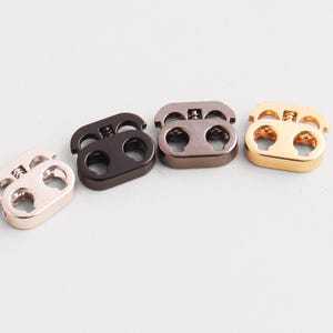 May include: Four metal cord locks in various colors: rose gold, dark brown, gray, and gold. Each cord lock has two holes and a spring mechanism for securing cords or drawstrings. These are commonly used in apparel and accessories.