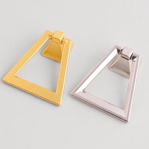 May include: Two metal drawer pulls. One is gold-toned, the other is silver-toned. Both have a triangular design with a rectangular base. These pulls are likely for cabinets or drawers, offering a modern aesthetic.
