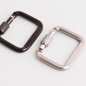 Könnte beinhalten: Zwei quadratische Karabinerhaken. Einer ist schwarz, der andere silberfarben. Beide haben einen Schraubverschlussmechanismus. Die Haken sind zum Sichern von Gegenständen konzipiert und werden häufig bei Outdoor-Aktivitäten verwendet.