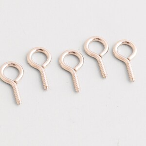May include: Five rose gold screw eye hooks arranged diagonally. Each hook features a closed loop and a threaded screw base. Ideal for crafts, jewelry making, or hanging small items. The hooks are made of metal.